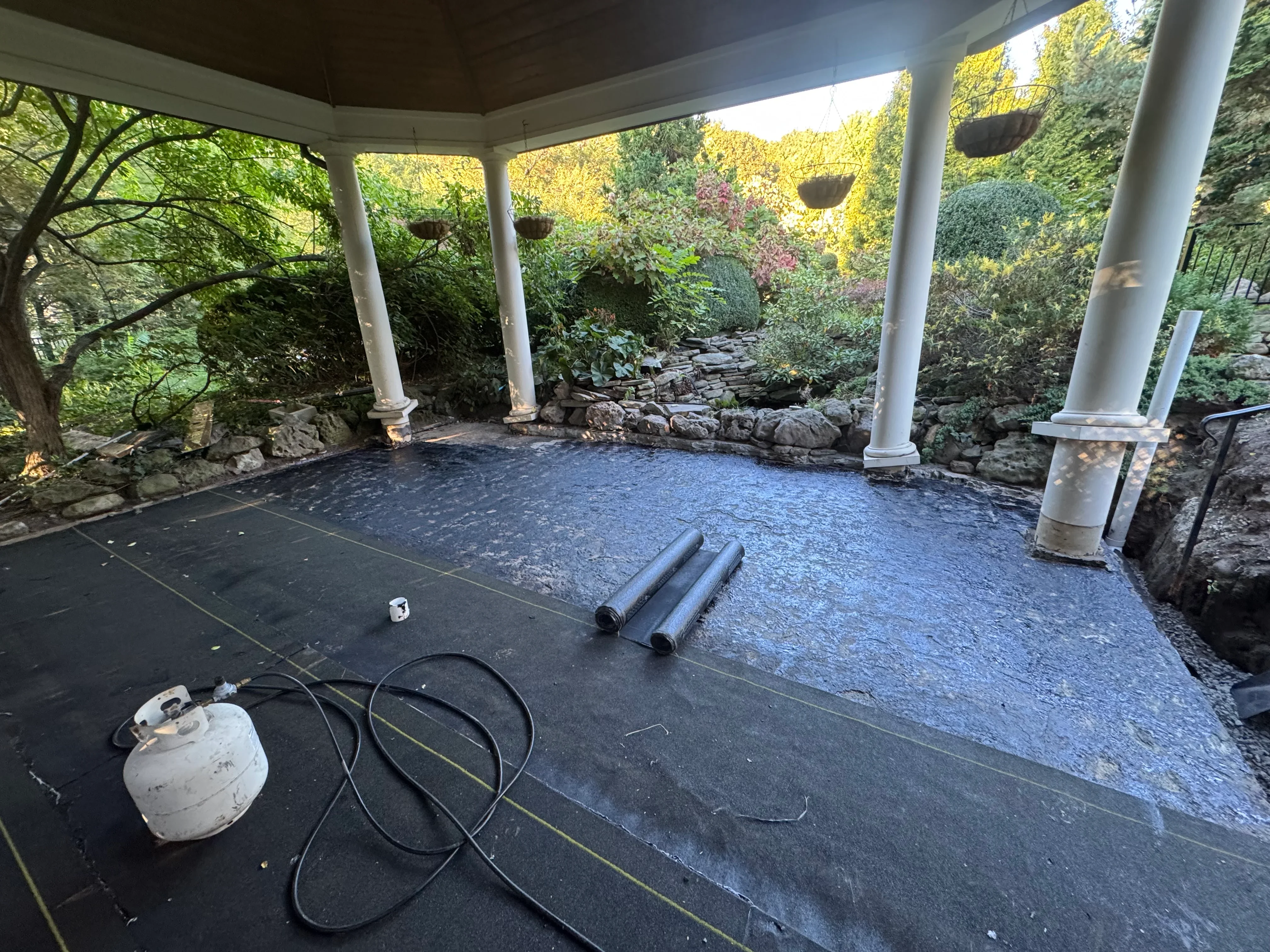 Covered patio with new dark roofing membrane, white columns, stone walls, and drainage installation in progress