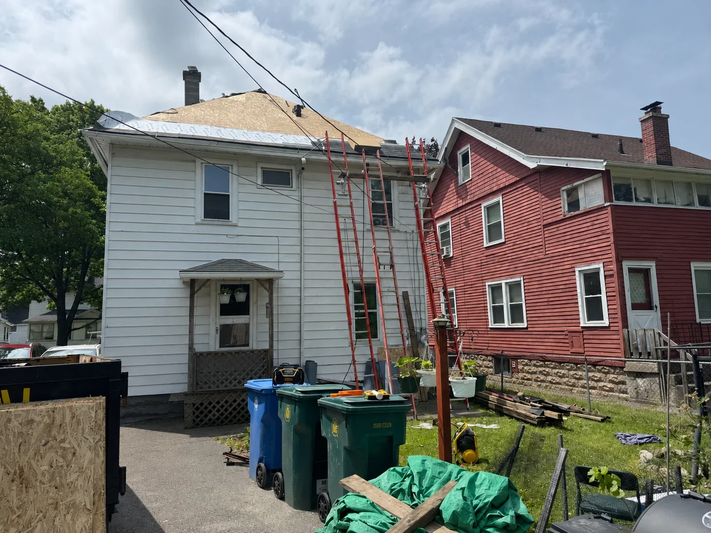 Two-story house with new roof framing and asphalt shingles during installation, ladders and materials visible